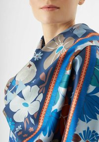 Foulard en soie avec un motif floral bleu, blanc et marron, bordé de rouge et de bleu ; texture douce, drapé élégamment.