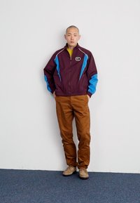 Jachetă vânt din material roșu-maroniu și albastru, cu fermoar și logo, asortată cu pantaloni maroni și pantofi bej. Textură netedă, design atletico.