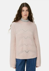 Eine Frau mit langen, welligen Haaren, die einen hellrosa Strickpullover über einem grauen Rollkragenpullover und blauen Jeans trägt und mit dem Gesicht zur Kamera schaut, vor einem weißen Hintergrund.