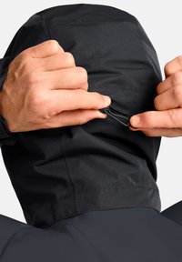 Veste imperméable noire avec capuche ajustable. Les mains tirent sur le cordon au niveau du cou pour un ajustement près du corps. Texture de tissu lisse, design minimaliste.