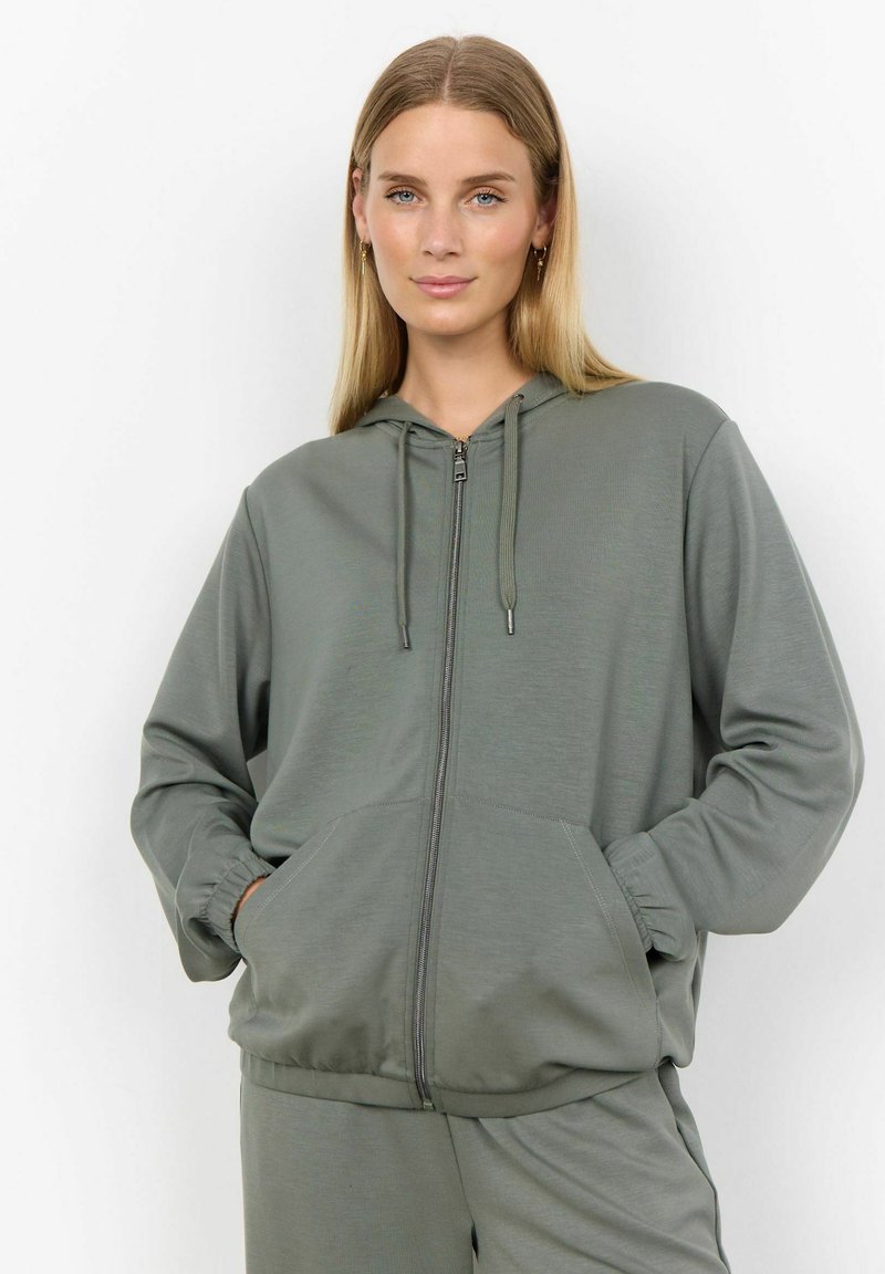 Khaki mikina na zips v mäkkom, štrukturovanom materiáli. Obsahuje kapucňu s šnúrkou, klokanie vrecká a elastické manžety. Nepretržitý a pohodlný dizajn.