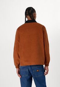 Brown corduroy jacket with a black collar, featuring long sleeves, a relaxed fit, and elastic cuffs. Denim jeans visible at the bottom.