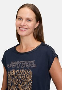 Navy blue t-shirt with short sleeves, features "JOYFUL" in rhinestones and a textured golden pattern, finished with a shimmering neckline.