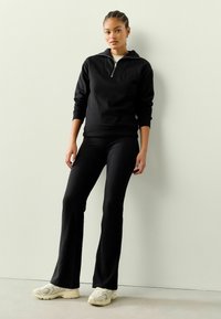Schwarze Zip-Up-Sweatshirt mit gerippten Bündchen, kombiniert mit schwarzen ausgestellten Leggings und klobigen weißen Sneakers, vor einem neutralen Hintergrund.