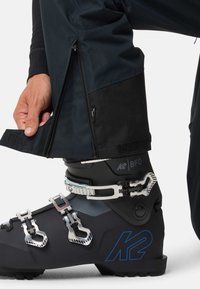 K2 skischchoenen in zwart met zilverkleurige hardware, voorzien van drie metalen gespen en een gestructureerde zool, gecombineerd met donkere buitenkleding met een ritsopening bij de benen.