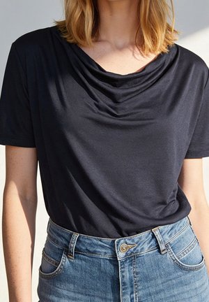 Femme portant un haut noir à manches courtes avec un col drapé, rentré dans un jean bleu taille haute avec des poches avant et une fermeture par bouton.