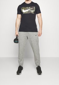 Camiseta negra de Nike con un gráfico de logo en camuflaje, combinada con pantalones de chándal gris claro y zapatos deportivos negros; sosteniendo un kettlebell negro.