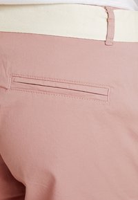 Pantalon rose clair avec une ceinture texturée beige, présentant une poche de couture horizontale à l'arrière. Tissu lisse avec une finition mate.