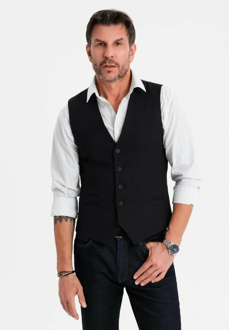 Mann mit kurzem dunklem Haar und Bart, der eine schwarze Weste über einem gestreiften weißen Hemd und dunklen Jeans trägt, steht mit der Hand in der Tasche.