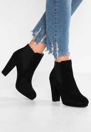 Bottines en daim noir à talons épais portées avec un jean bleu à ourlet effiloché sur un sol blanc.