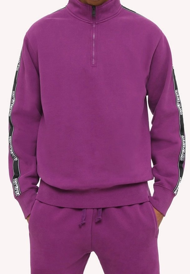 Personne portant un sweat-shirt violet à fermeture éclair au quart et un pantalon violet assorti avec des détails de bande rayée noire et blanche sur les manches.