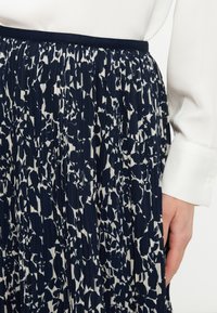 Πλισέ φούστα με ναυτικό μπλε και λευκό αφηρημένο λουλουδάτο μοτίβο, φορεμένη με λευκό πουκάμισο μακρυμάνικο, μανσέτα ορατή δίπλα στο χέρι.