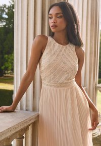 O rochie fără mâneci, de lungime până la podea, în nuanțe delicate de roz, cu un fustă plisată. Bustul are mărgele intricate aranjate într-un model diagonal.