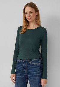 Langarmshirt aus dunkelgrünem Rippstoff, runder Ausschnitt, mit kleinem Logo auf der Brust, kombiniert mit blauen Jeans.