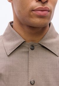 Άνδρας που φοράει μπεζ πουκάμισο με γιακά και μαύρα κουμπιά κλιπς, με εμφανές το κάτω μέρος του προσώπου και τον λαιμό, σε απλό φόντο.