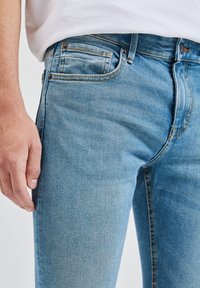 Světle modré džíny z denimu s úzkým střihem, předními kapsami a kontrastními švy. Tkanina má hladkou texturu a mírnou pružnost.