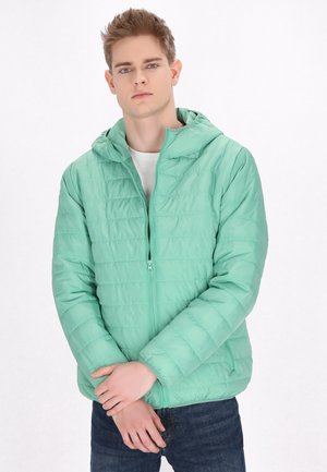 Jachetă puf cu glugă de un verde deschis, cu un design matlasat, față cu fermoar și manșete elastice, asortată cu blugi negri și o cămașă albă.