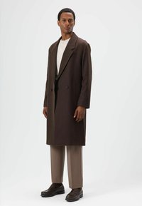 Bruine dubbelgeknoopte wollen jas met brede revers, zijzakken en een halflange snit, gecombineerd met lichtbeige broek en bruine loafers.