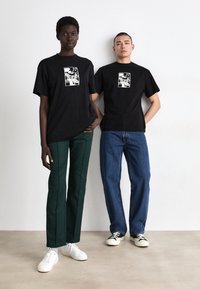 Dva posameznika stojita drug ob drugem. Nosita črne majice s grafičnim dizajnom, v kombinaciji z zelenimi in modrimi hlačami ter belimi supergami.