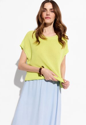 Kobieta o falowanych brązowych włosach, ubrana w limonkowy krótki sweter z krótkim rękawem, jasnoniebieską spódnicę i bransoletkę z motywem muszli żółwia, trzymająca brzeg swojego topu.