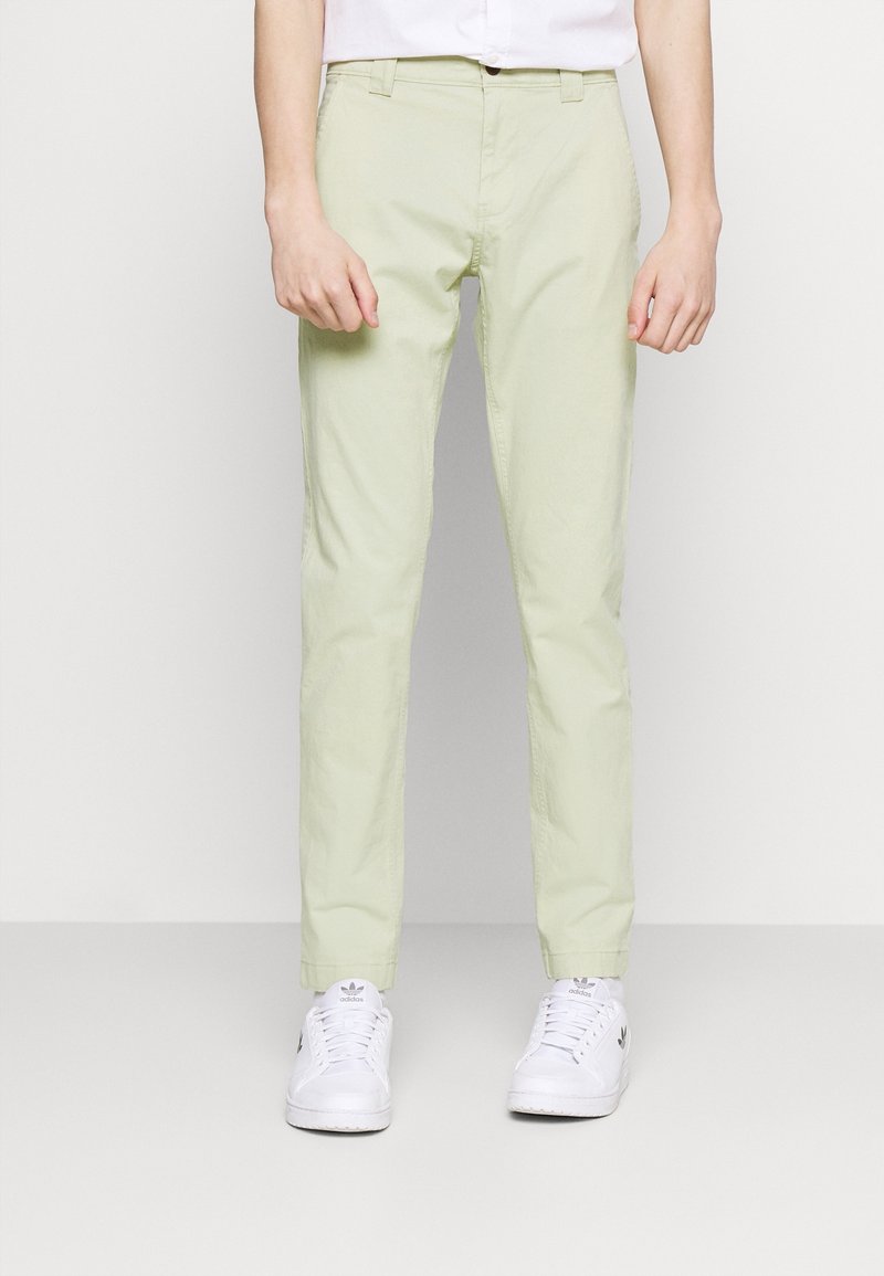 Personne portant un pantalon droit vert clair et des baskets Adidas blanches, debout devant un fond blanc uni.