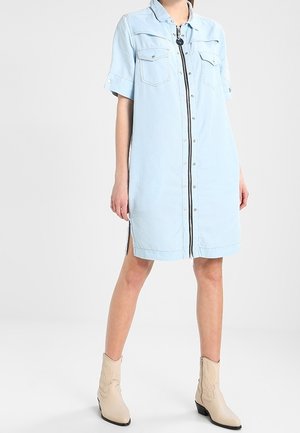 Kobieta w jasnoniebieskiej sukience koszulowej z denimu z zamkiem z przodu oraz beżowymi botkami, stojąca na tle jasnego, jednolitego tła.