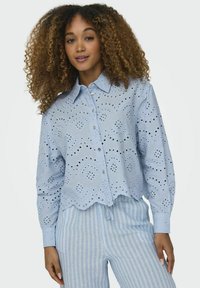 Camisa de botones ligera de color azul con bordado de ojales, dobladillo en forma de onda y mangas largas. Combinada con pantalones rayados de color azul claro.