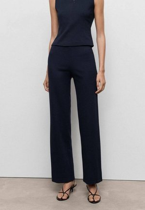 Femme portant un haut sans manches bleu marine, un pantalon large assorti et des sandales noires à talons à brides, debout contre un mur clair uni.