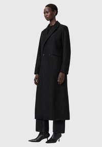 Abrigo largo negro con diseño de doble botonadura, ajuste entallado y solapas muescadas. Llevado sobre jeans oscuros y tacones puntiagudos. Textura de tela suave.