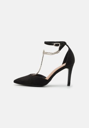 Black suede high heel shoe with pointed toe, ankle strap, and chain detail connecting strap to shoe. Glossy heel, sleek design.