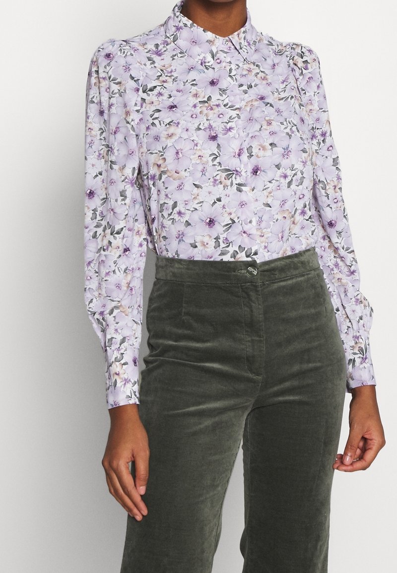 Femme portant un chemisier lilas à motif floral à manches longues, rentré dans un pantalon en velours vert foncé taille haute, debout devant un fond blanc.