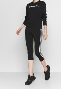 Zwart sweatshirt met wit "Champion" logo, gecombineerd met zwarte caprileggings met witte strepen aan de zijkant. Model draagt zwarte sportschoenen.