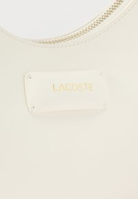 Λευκό δερμάτινο αξεσουάρ με χρυσό φερμουάρ και ραμμένο ορθογώνιο πατς με την επιγραφή "LACOSTE" σε χρυσά γράμματα.