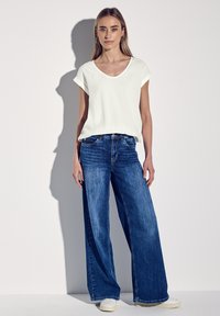 Bílý top s krátkým rukávem a volným střihem, kombinovaný s volnými modrými džínami. Modelka stojí na jednoduchém pozadí.