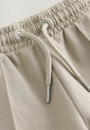 Bézs színű melegítőnadrág elasztikus derékrésszel, ezüst szemekkel és bézs húzózsinórokkal. Puha pamut keverék anyag, sima textúrával.