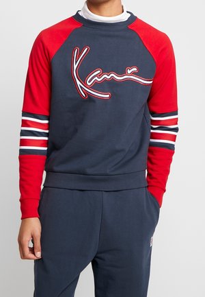 Man draagt een marineblauwe sweatshirt met rode raglanmouwen en gestreepte details, met een grote "Kani"-logo op de borst.