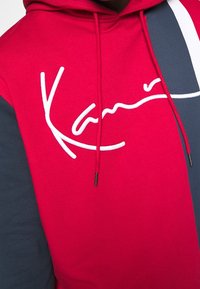 Sudadera roja con bloques de color azul marino y blanco en las mangas, con texto blanco bordado en estilo firma en el pecho y cordones rojos.