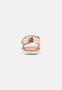 Sandály v růžově zlaté metalické barvě s minimalistickým designem, tenkými pásky a bílou podrážkou. Vnitřní část se zdá být texturovaná v hnědé barvě.