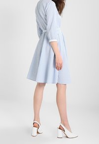 Kvinna som bär en ljusblå rutmönstrad klänning i knälängd med trekvartsärmar och vita klackslippers.