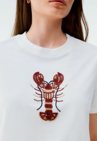 Tricou alb cu un motif de homar, având mărgele roșii, negre și aurii. Mâneci scurte și decolteu ribatat. Textură netedă de bumbac.