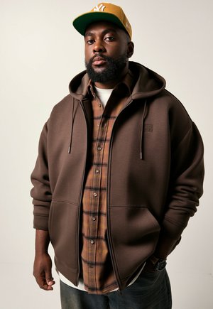 Man with beard wearing mustard cap, brown zip-up hoodie, plaid shirt, white undershirt, jeans, and wristwatch against plain background.