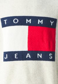 Sudadera con un logo de punto que dice "TOMMY" en azul marino sobre un fondo crema y un diseño rectangular en rojo y blanco debajo.