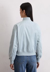 Kobieta z długimi ciemnymi włosami, ubrana w jasnoniebieską kurtkę bomberkę i niebieskie dżinsy, stoi twarzą do gładkiej, jasnoszarej ściany.