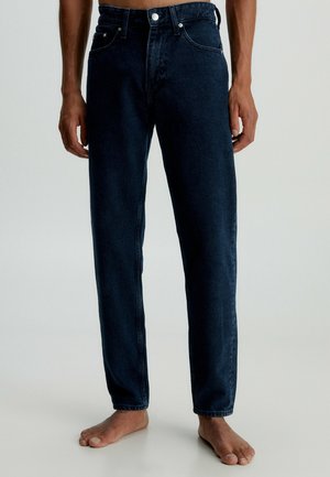 Barfüßige Person, die dunkelblaue Jeans mit geradem Bein trägt und vor einem einfarbigen hellen Hintergrund steht.