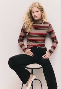 Brūnas, sarkanas un melnas svītrainas augstu aizvērtas apkakles tops kopā ar melnām izplatītām biksēm un baltām kurpēm, sēžot uz krēsla pret neitrālu fona.