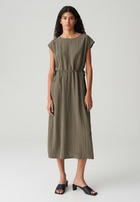 Olivenfarbene knielange Kleid mit kurzen Ärmeln, gerafftem Taillenband und strukturiertem Stoff. Kombiniert mit schwarzen Slippern mit Blockabsatz.