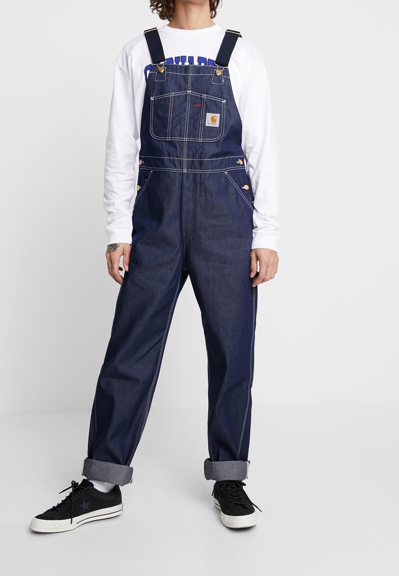 Jeansoverall i mörkblått med justerbara axelremmar, framficka och detaljer i mässing. Upprullade ärmslut med en avslappnad passform.