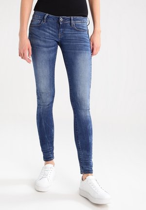 Jeans ajustados en denim azul medio, con un diseño clásico de cinco bolsillos, un ligero desvanecimiento y un corte ajustado. Combinados con zapatillas blancas.