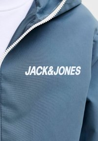 Veste bleue avec fermeture éclair blanche partiellement ouverte, montrant le logo "JACK & JONES" en texte blanc sur la poitrine.