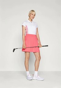 Moteris stovi laikydama golfo lazdelę horizontaliai, vilkinti baltą polo marškinėlį su rožinėmis gėlėmis šonuose, rožinę plisėtą sijoną, baltas kojines ir baltus sportinius batelius.